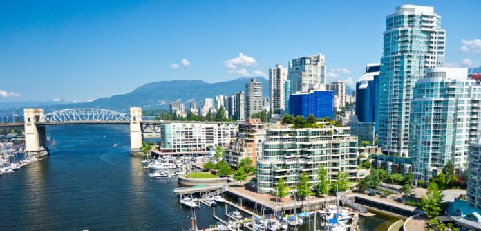 Aerial view of a coastal city with tall modern buildings, a bridge over a body of water, and docks with boats on a sunny day. Mountains in the background reflect the vibrant atmosphere captured in BC’s Quarterly Market Overview.