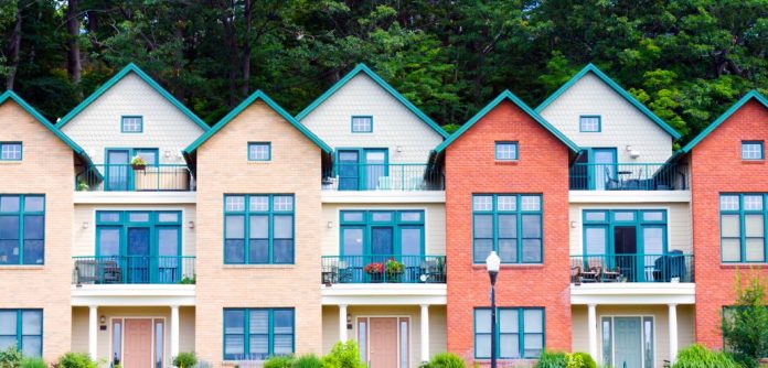 Row of colorful townhouses with brick and beige facades, peaked roofs, and green trim, surrounded by trees and greenery. The buildings have large windows and balconies.