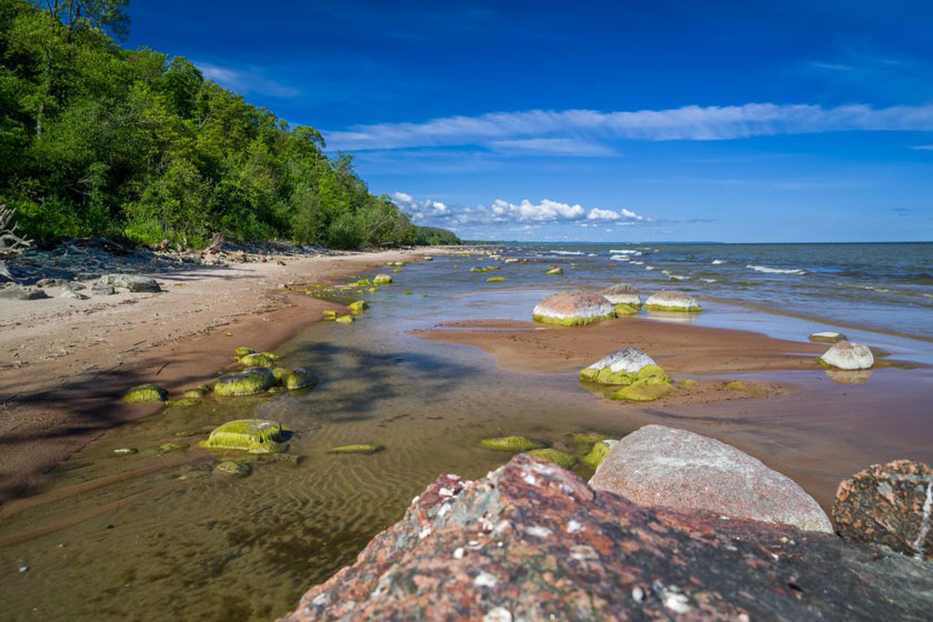 A sandy shore with scattered moss-covered rocks, clear shallow water, and a forested area on the left, under a blue sky with scattered clouds.