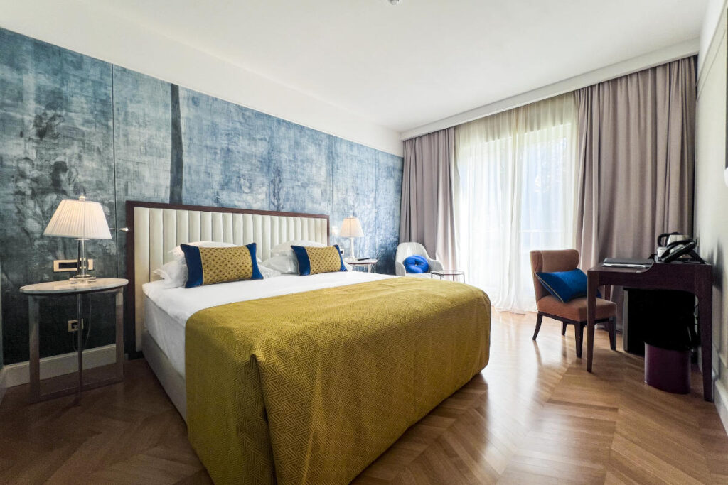 A Hotel Milenij room with a large bed featuring yellow bedding, two lamps on bedside tables, a desk with a chair, and a sitting area near a window with curtains. Blue accent pillows adorn the bed and chair.