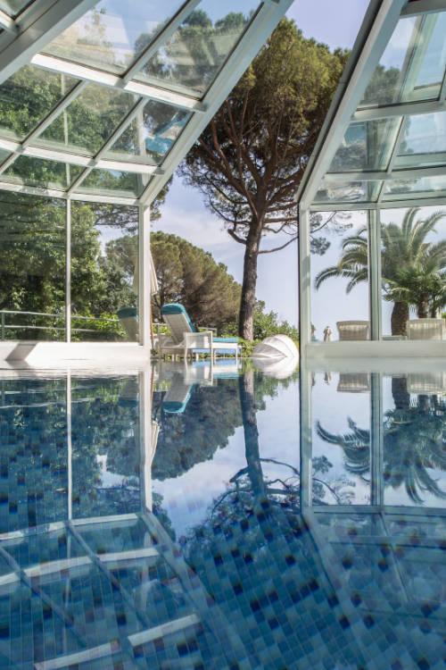 The indoor swimming pool at Hotel Milenij features glass walls and a ceiling, offering views of a large tree and palm trees outside. Lounge chairs and a small table can be seen through the glass, with the water reflecting the picturesque surroundings.