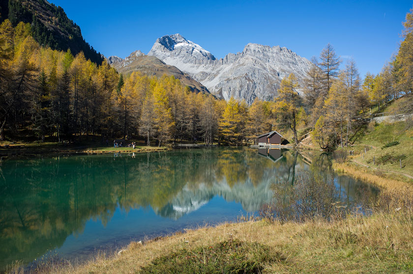 A tranquil lake with clear water reflects snow-capped mountains, autumn trees, and a wooden cabin on the shore under a clear blue sky.