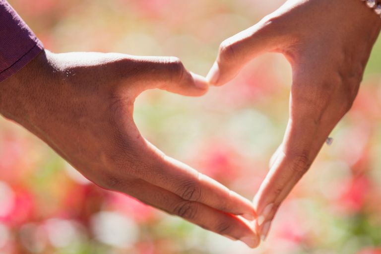 two hands forming a heart shape against a blurred floral background