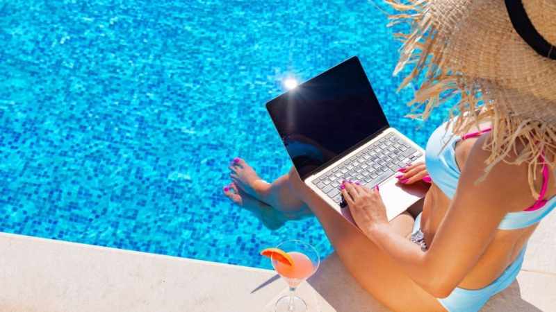 workation with hotelsbyday: A person in a sun hat and swimsuit works on a laptop by a vibrant blue pool, with a cocktail on the side.