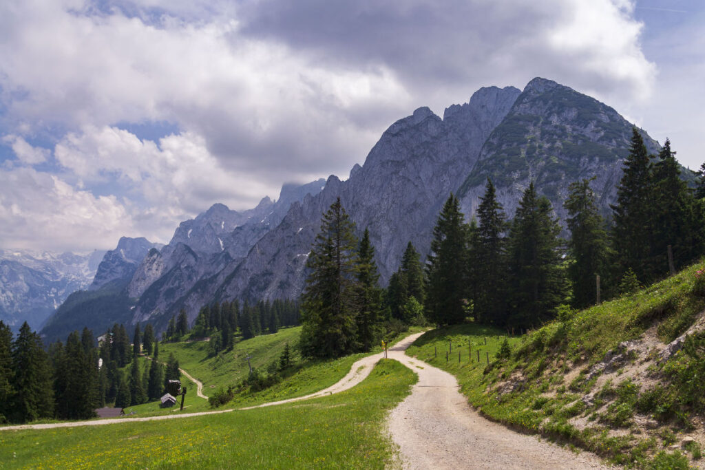 A winding dirt path leads through a grassy landscape with scattered trees, set against a backdrop of tall, rugged mountains under a partly cloudy sky, marking it as one of the best places to visit for nature enthusiasts seeking breathtaking views.