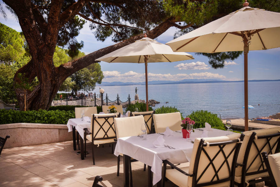 The outdoor dining area at Hotel Milenij features tables and chairs under umbrellas on a charming patio, overlooking a scenic body of water with trees in the background.