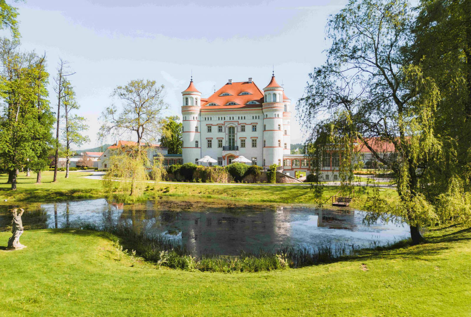 A grand white building with red rooftops, surrounded by trees and greenery, with a pond in the foreground on a clear, sunny day, reminiscent of the stunning Pałac Wojanów.