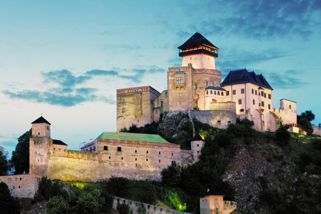 A large medieval castle with towers and walls sits atop a hill against a sunset sky.