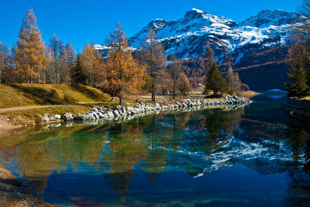 Scenic view of a lake with rocky shoreline, surrounded by autumn trees and snow-capped mountains reflecting in the clear water under a bright blue sky.